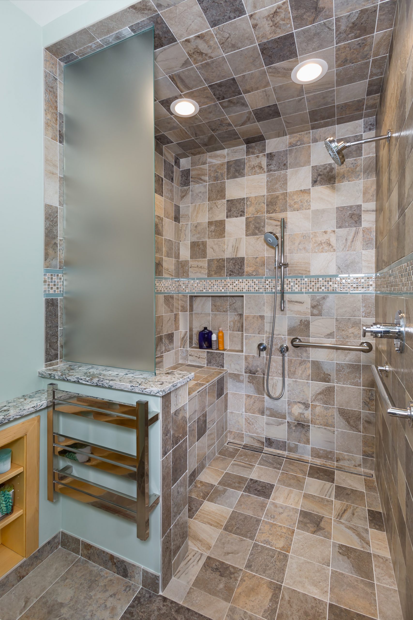Walk in shower with bright lighting, light brown custom square tile, removable spray head, shower seating and grab bars.