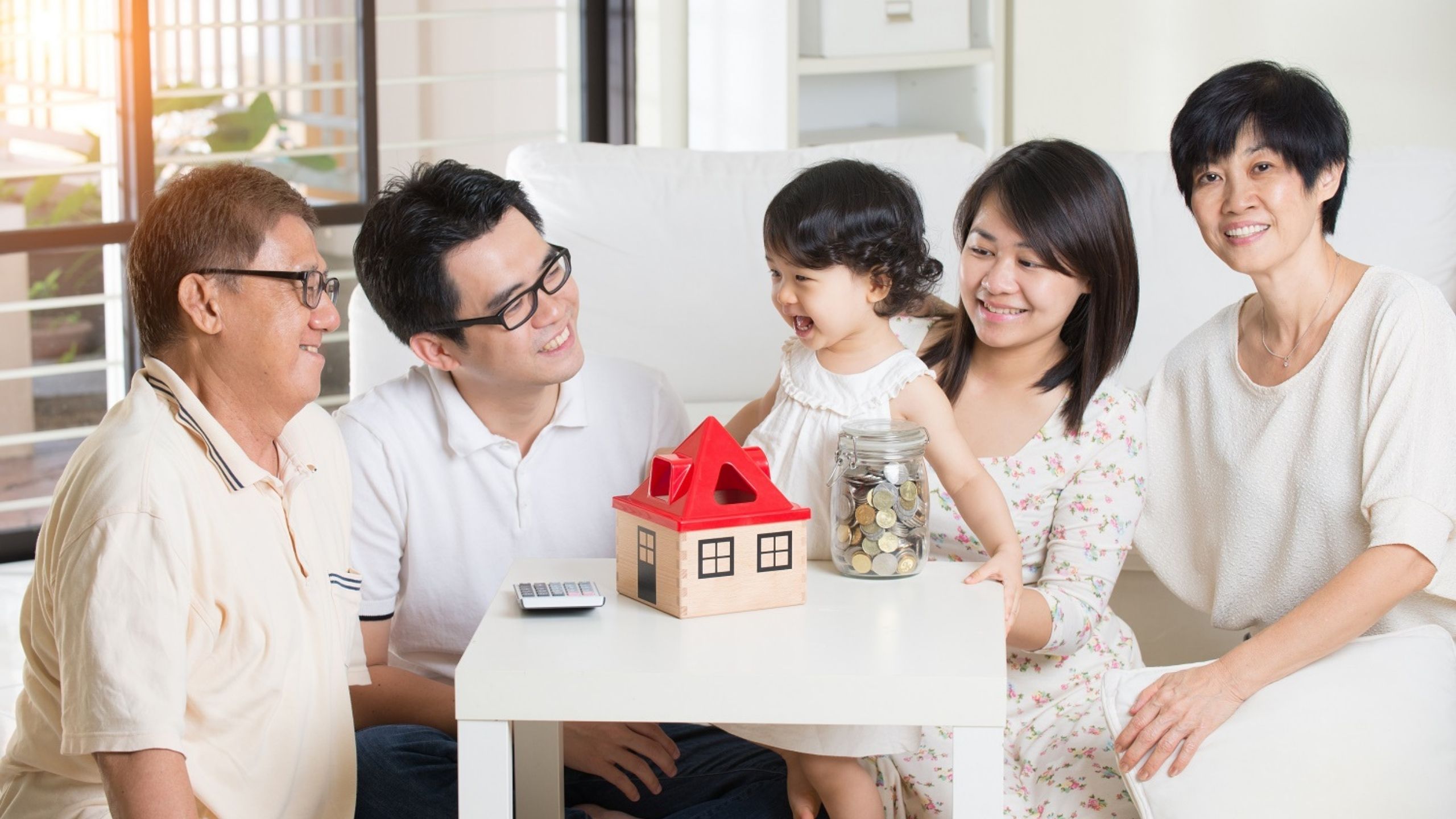 Three generation family smiling together with small toy house and jar of coins on table
